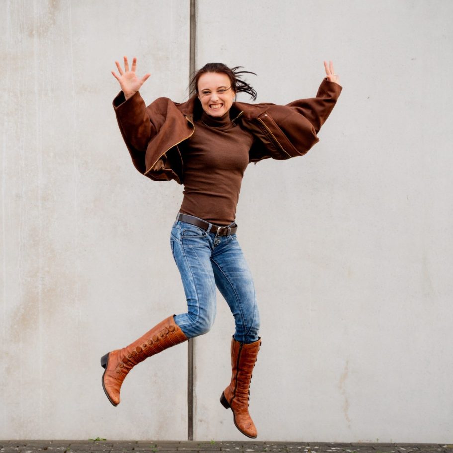 Eine lachende Frau springt fröhlich in Jeans und braunen Stiefeln vor einer grauen Wand.